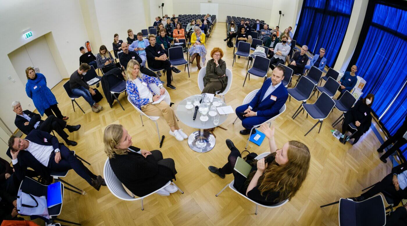 Teilnehmer der 21. Jahreskonferenz des RNE sitzen in der Mitte eines Raumes, umringt von Zuschauern, und sprechen miteinander über das Thema nachhaltige Innovationen. © André Wagenzik/Florian Bolk, RNE