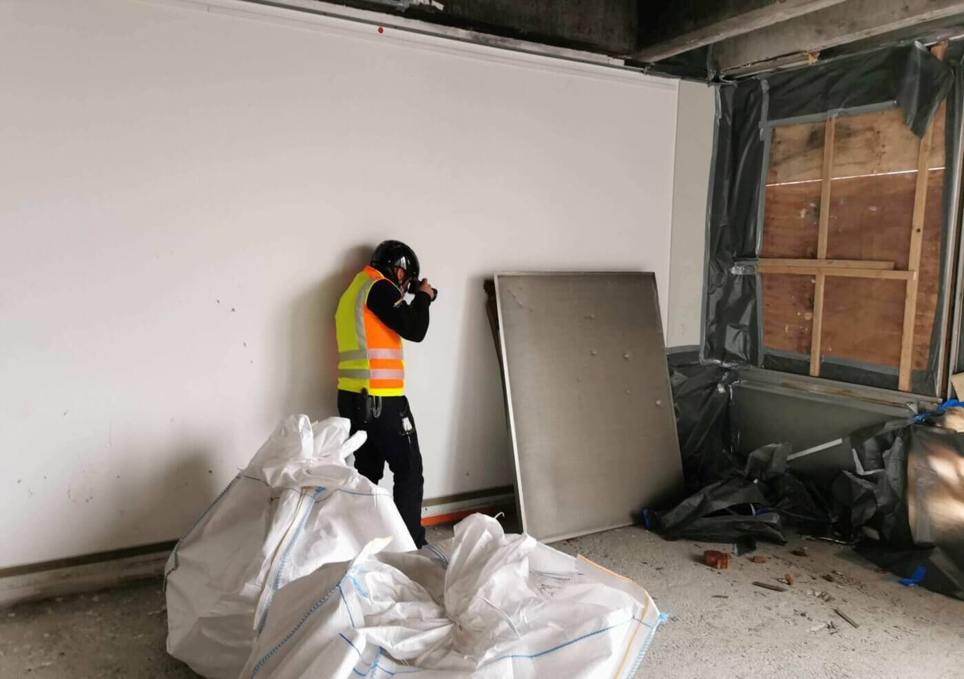 Ein Mann mit Sicherheitsweste und Helm fotografiert auf einer Baustelle in einem Raum mit vernageltem Fenster und Bauschutt.