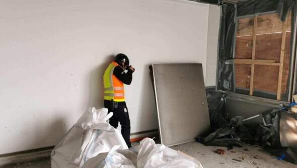 Ein Mann mit Sicherheitsweste und Helm fotografiert auf einer Baustelle in einem Raum mit vernageltem Fenster und Bauschutt.