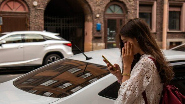 Eine Frau steht an einer Straße vor Autos und schaut auf ein Handy.