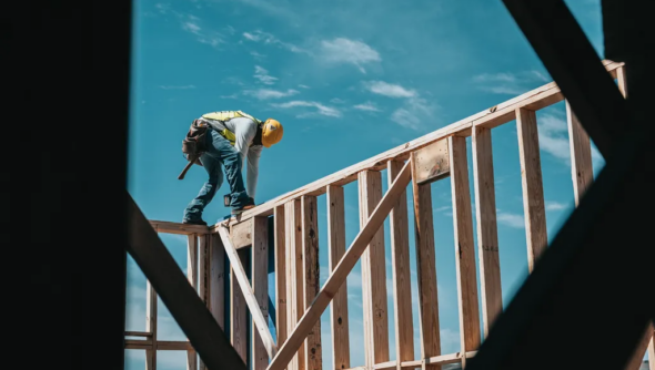 Ein Bauarbeiter balanciert gebückt auf einem Holzgerüst und bearbeitet das Holz. Im Hintergrund sieht man blauen Himmel.