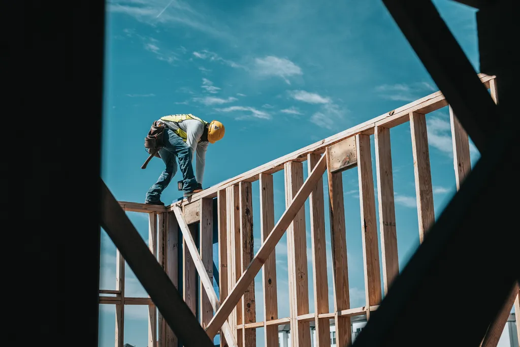Ein Bauarbeiter balanciert gebückt auf einem Holzgerüst und bearbeitet das Holz. Im Hintergrund sieht man blauen Himmel.