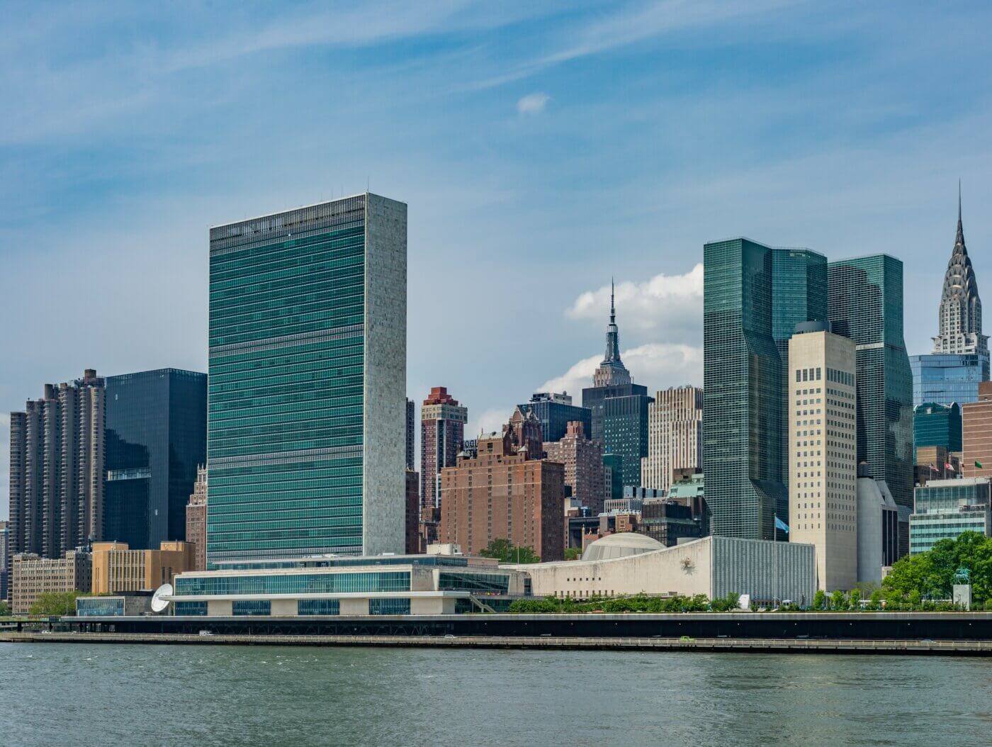 Auf dem Bild sieht man die Skyline von New York vom Wasser aus mit blauem Himmel im Hintergrund. Im Vordergrund steht das hohe und eckige Gebäude der Vereinten Nationen UN.