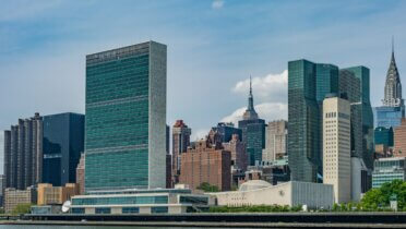 Auf dem Bild sieht man die Skyline von New York vom Wasser aus mit blauem Himmel im Hintergrund. Im Vordergrund steht das hohe und eckige Gebäude der Vereinten Nationen UN.