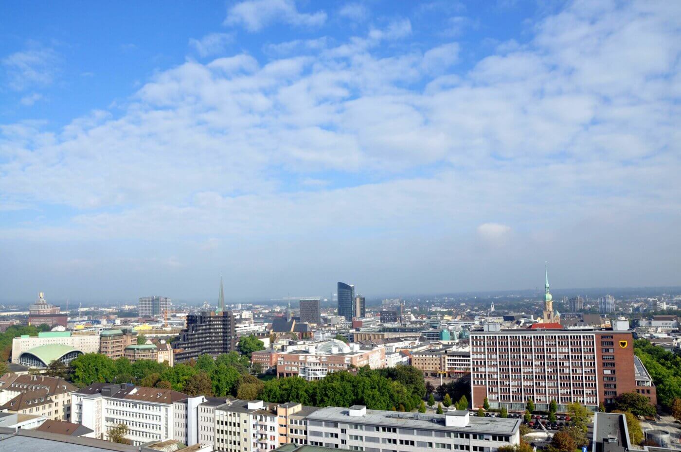 Ein Panoramablick von oben auf die Stadt Dortmund. In der Mitte sieht man ein Hochhaus und weiter rechts einen Kirchturm.