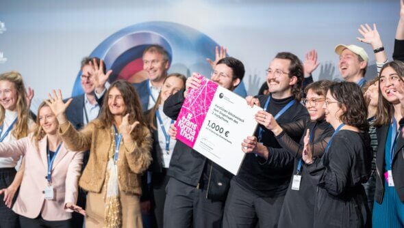 Die Gewinner von Wohnen+ stehen auf der Bühne bei der Verleihung des Deutschen Nachhaltigkeitspreises und halten ein Schild mit dem Preisgeld hoch.