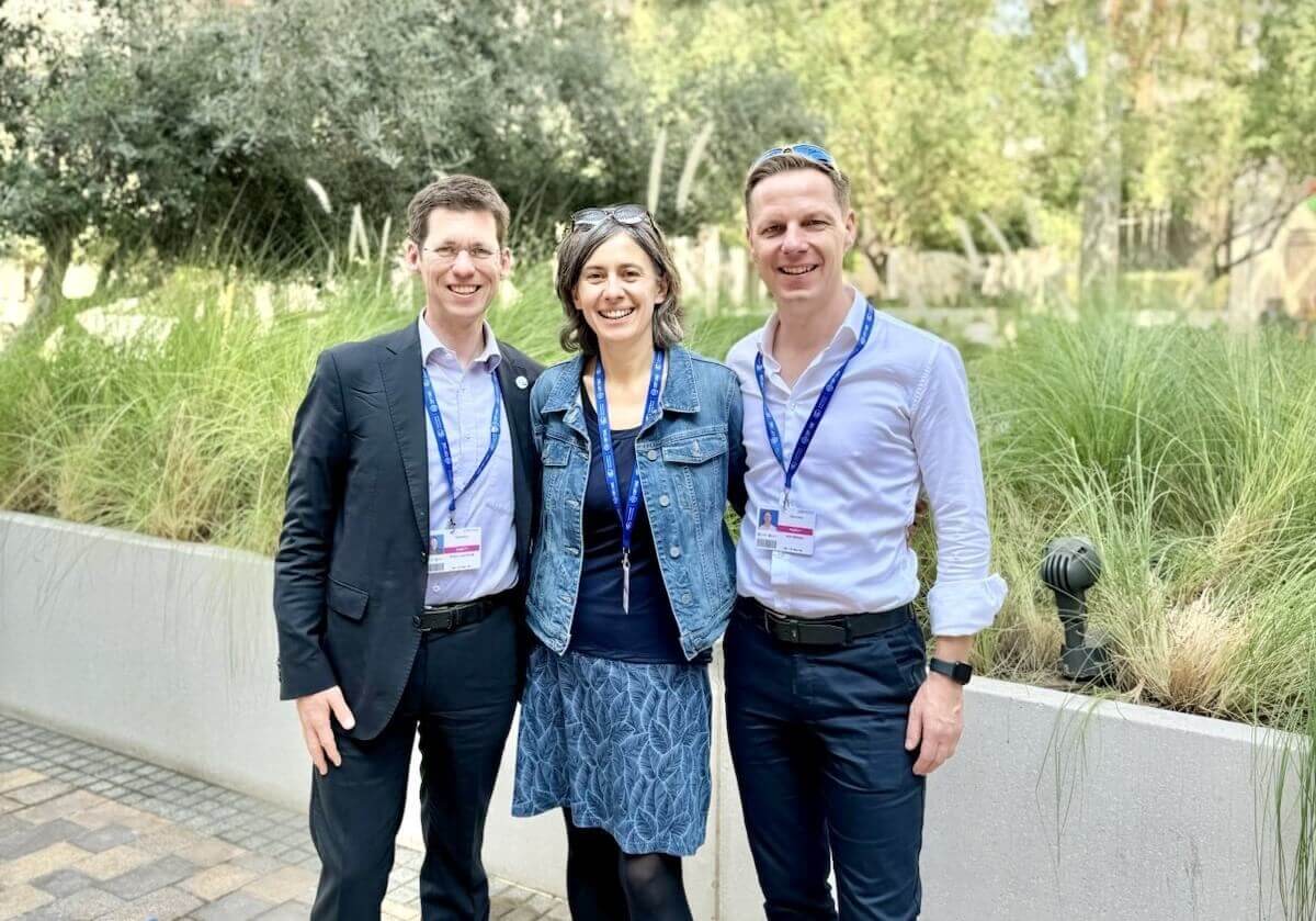 Die Ratsmitglieder Mark Lawrence, Franziska Tanneberger, Kai Niebert (von links nach rechts) stehen vor einer umrandeten grünen Wiese mit Gräsern und Bäumen in Dubai. Foto: RNE