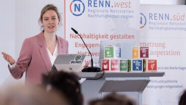 Myriam Rapior, stellvertretende Bundesvorsitzende des BUND e.V. und RNE-Mitglied hält einen Vortrag auf den RENN Tagen 2023. Neben ihr sind Banner der RENN sowie SDG Würfel zu sehen.