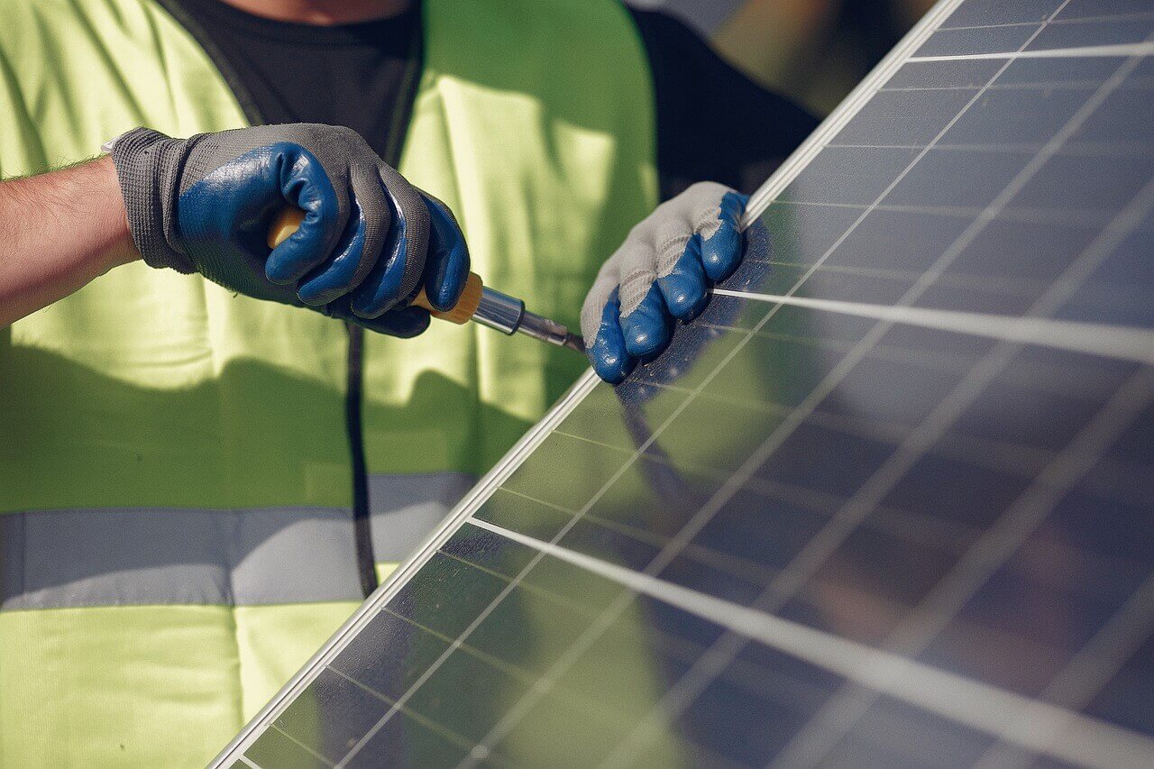 Auf dem Foto sieht man den einen Teil eines Solarpanel. Am Rand steht ein Arbeiter mit Handschuhen und einer gelben Weste und schraubt mit einem Schraubenzieher eine Schraube fest.