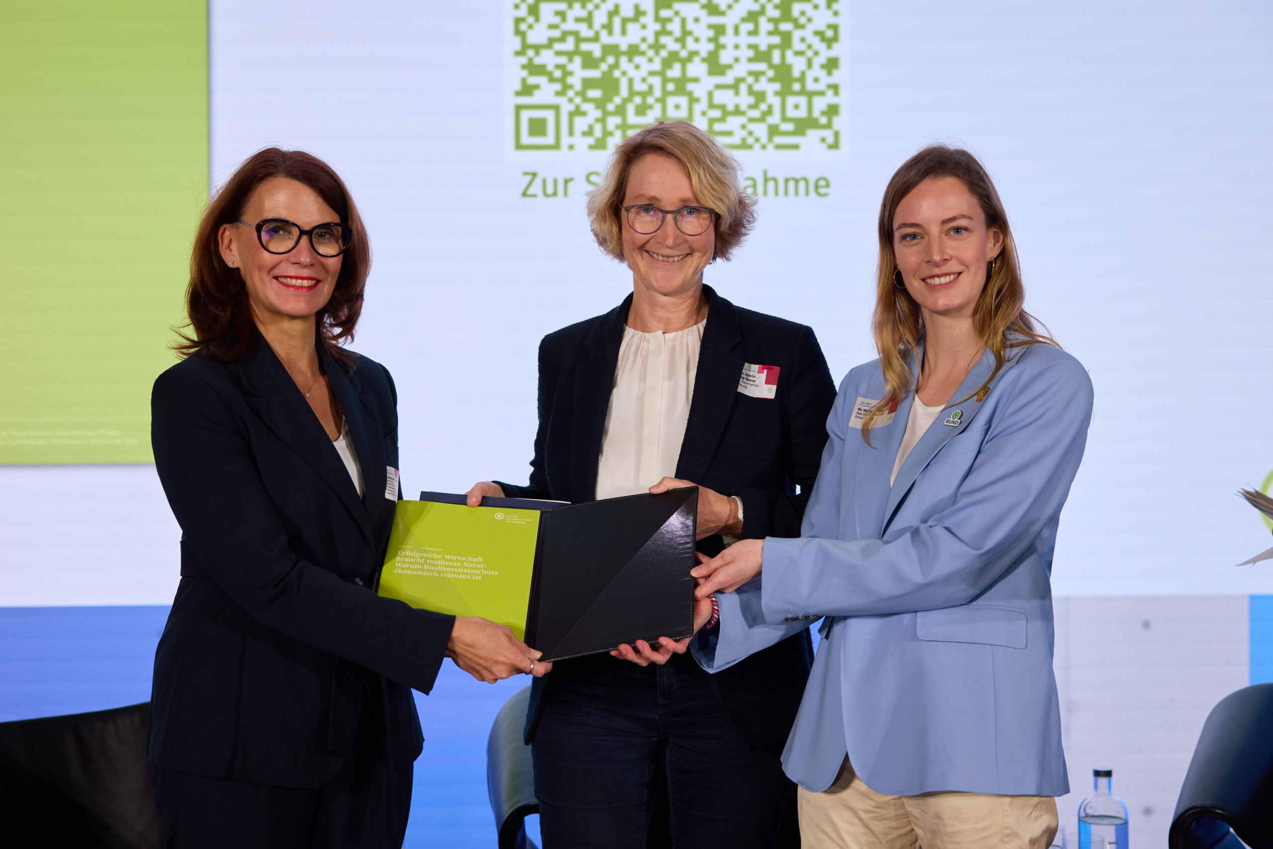 Drei Frauen stehen auf einer Bühne und halten eine Dokumentenmappe mit einer Stellungnahme in der Hand.