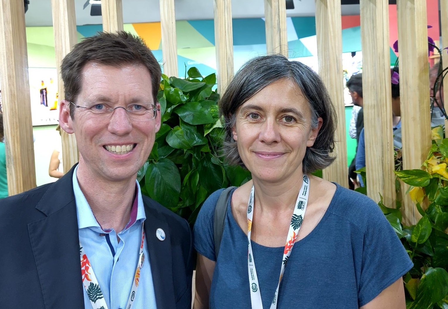 Die Ratsmitglieder Franziska Tannenberger und Mark Lawrence stehen vor dem deutschen Pavillon bei der COP30 in Brasilien. Im Hintergrund sieht man eine Trennwand aus Holz und Pflanzen sowie einen bunten Hintergrund.