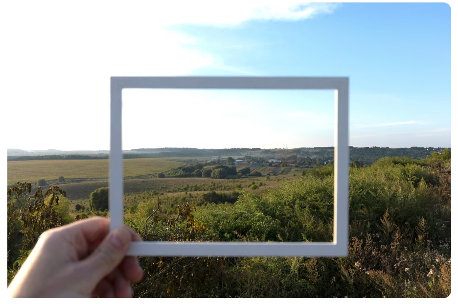 Auf dem Foto sieht man eine hügelige, grüne Landschaft mit Büschen und in der Ferne ein paar kleine Häuser. Im Vordergrund hält eine Hand einen weißen Bilderrahmen, durch den man durchschauen kann und dahinter im Fokus die Landschaft sieht.