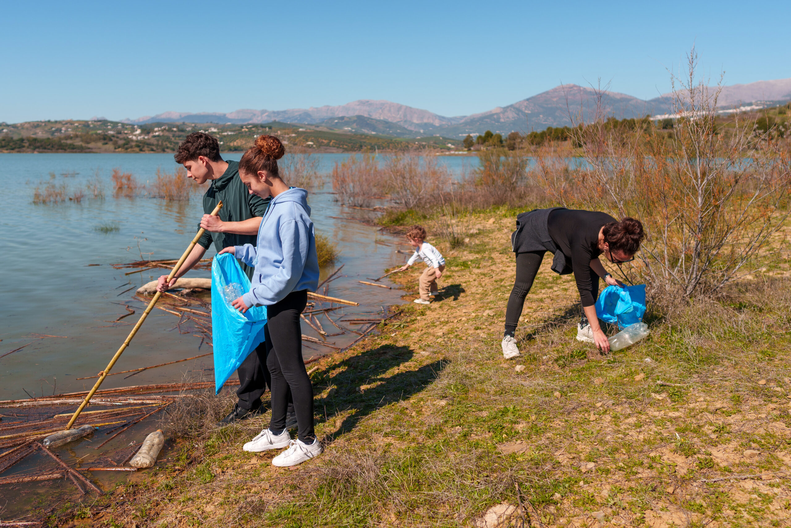 Eine Familie sammelt Plastikflaschen und Müll am Seeufer, vermittelt Umweltbewusstsein und ehrenamtliches Engagement. Im Hintergrund sind Berge am anderen Seeufer vor blauem Himmel zu sehen.
