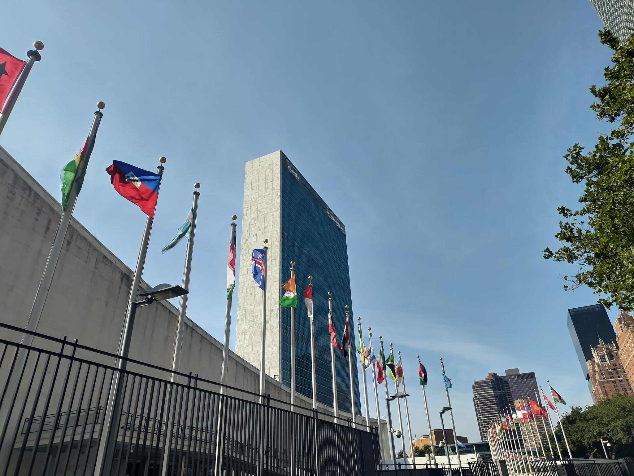 Hauptgebäude der Vereinten Nationen in New York vor blauem Himmel, im Vordergrund zahlreiche Flaggen.