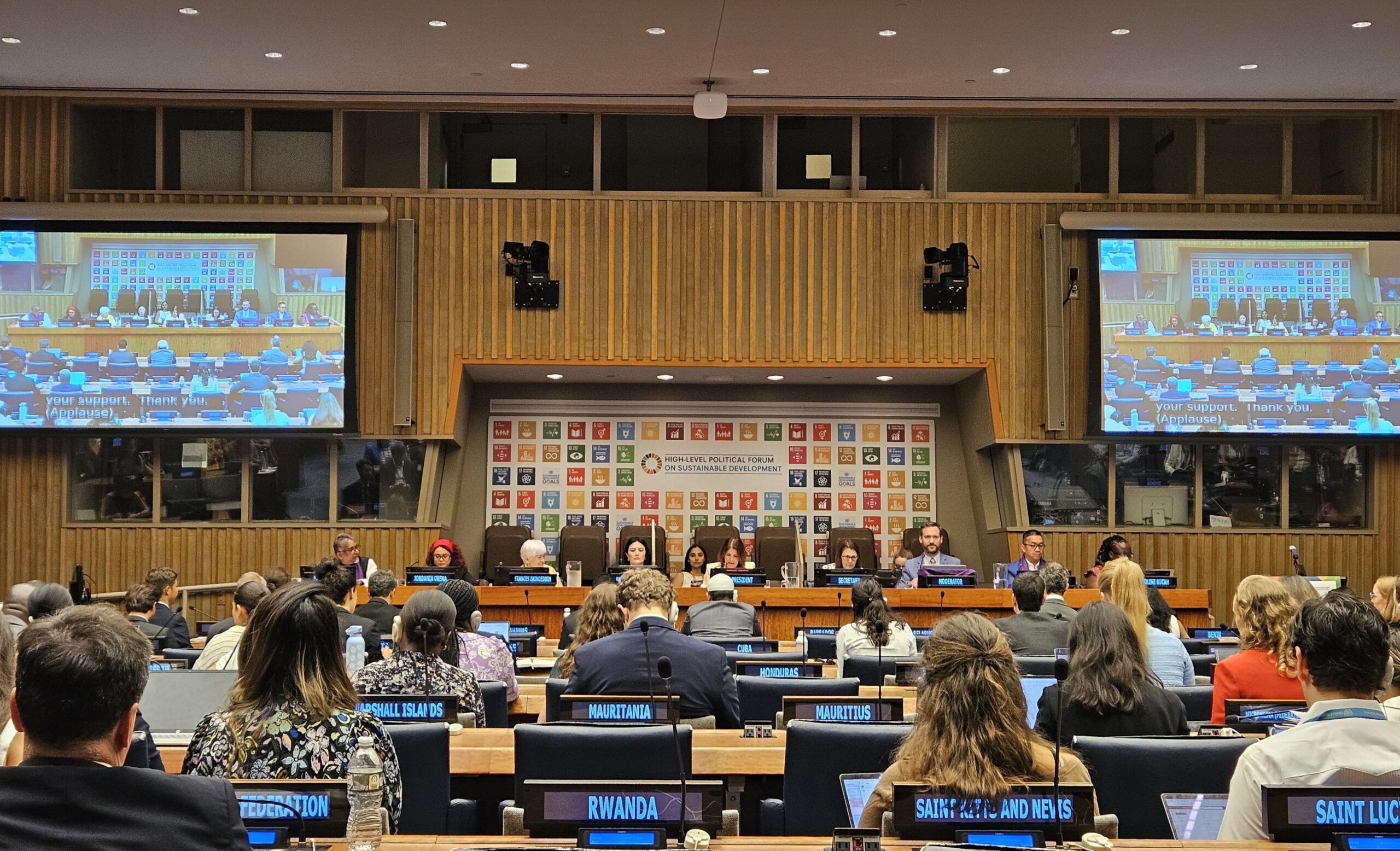 Auf dem Foto sieht man einen großen Raum im UN Gebäude in News York. Im Hintergrund sind auf einer Wand alle Symbolde der SDGs, eingerahmt von zwei großen Monitoren. Es findet gerade eine Versammlung statt, das heißt es sitzen Menschen auf der Bühne und im Publikum.